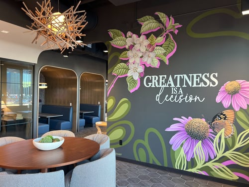 Breakroom with a brown table surrounded by several grey chairs. "Greatness is a decision" and flowers appear on the wall decorate the wall.