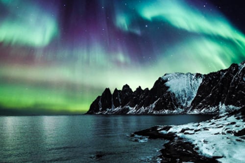 Northern lights dancing above snowy mountains and calm water under starry night sky.
