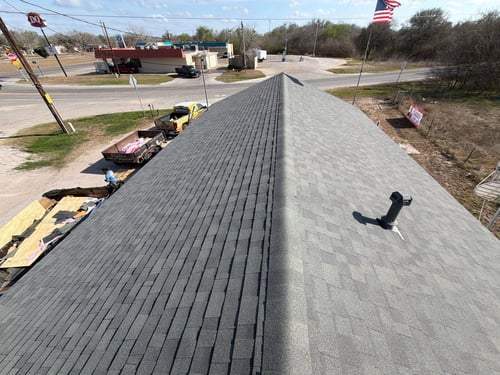 A newly shingled roof with a chimney vent runs symmetrically through the center. Below, workers and materials are visible. An American flag waves nearby.