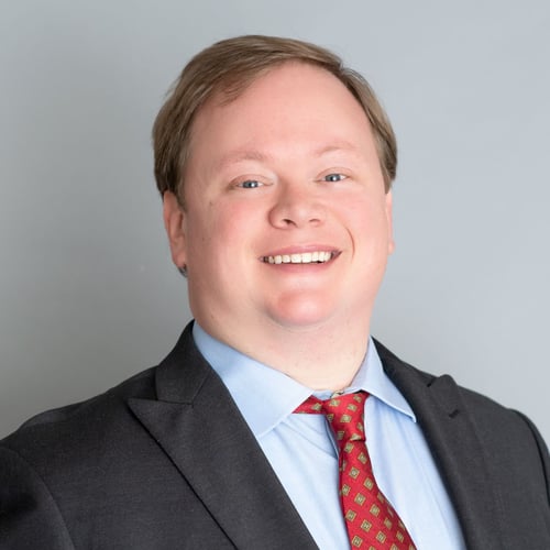 Agent Ryan standing and smiling wearing blue button up shirt with red tie and black blazer