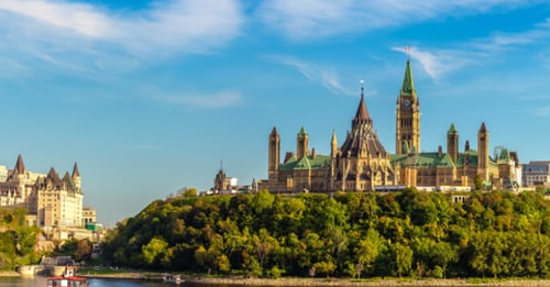 Gothic parliament buildings with clock tower on tree-covered hill above river under blue sky.