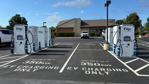 Several EVgo fast charging units installed in marked parking spaces at a public charging station, with EVgo logos on the chargers, protective bollards around each unit. Pull through parking spaces for charging. Storefront in the background.