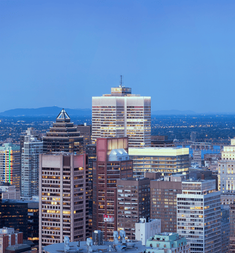 A city skyline at dusk featuring illuminated skyscrapers against a clear blue sky. The scene conveys a serene urban atmosphere.