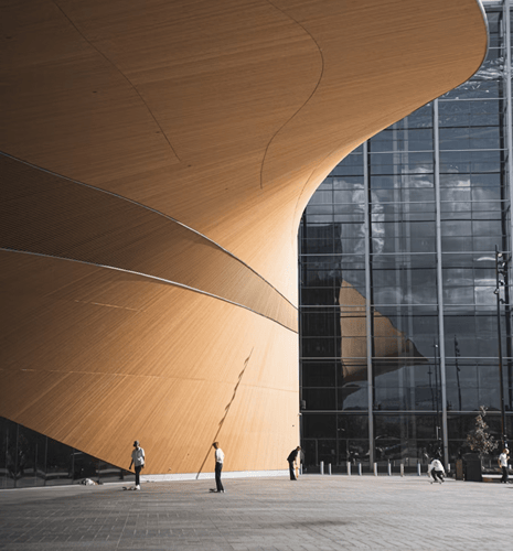 Modern architectural scene featuring a sweeping wooden curve against a glass facade, with people skateboarding. The mood is dynamic and urban.