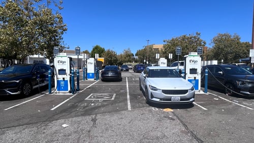 Several EVgo fast charging units installed in marked parking spaces at a public charging station, with EVgo logos on the chargers, protective bollards around each unit.