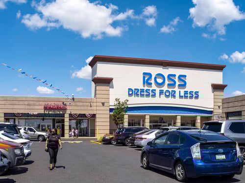 Woman walking through full parking lot as blue car passes by in front of Ross Dress for Less on a sunny day