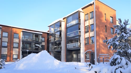 Ulkokuva Teeristä. Kerrostalorakennus on punertavaa tiiltä. Taivas on sininen ja aurinko paistaa. Maassa on runsaasti lunta pihan keskelle on kolattu korkea lumikasa.