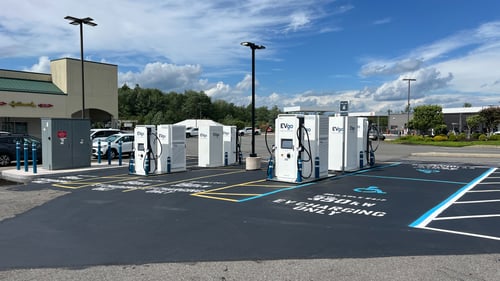 Several EVgo fast charging units installed in marked parking spaces at a public charging station, with EVgo logos on the chargers, protective bollards around each unit. Pull through and ADA stalls in image. Store fronts in background.