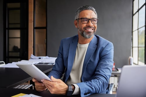 A man in a blue blazer and eyeglasses sits at a desk, holding a tablet. He smiles confidently, with an office backdrop and natural light streaming in.