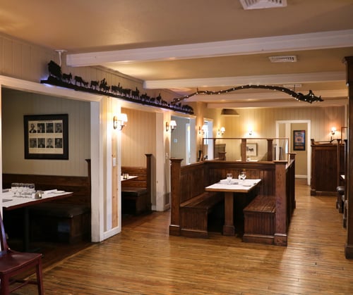 Interior of JK’s restaurant featuring classic wooden booths with white-clothed tables, warm wall sconces, and wood plank flooring. A decorative silhouette of a western cattle drive and string lights add rustic charm above the booths.