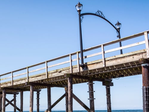Photograph of a wooden pier extending over calm ocean water under a clear blue sky.