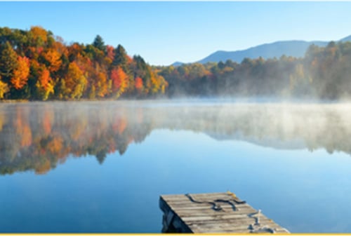 A tranquil lake reflects vibrant autumn trees and mist, with a wooden dock in the foreground. Mountains under a clear blue sky complete the serene scene.