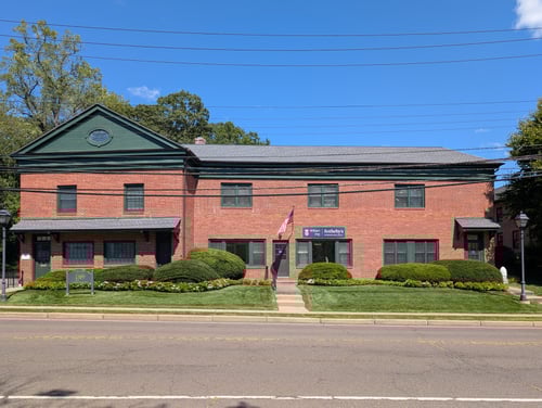 Two-story brick building with green trim and pediment featuring William Pitt Sotheby's International Realty on first floor right side in Southport (Fairfield), Connecticut, with American flag and manicured landscaping.