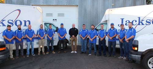 A group of 13 men in blue collared shirts stands in a line outdoors between two white vans with "Jackson" partially visible on the sides.
