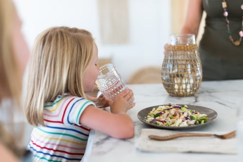 Child in a colorful striped top drinking water at a kitchen counter, watching a parent preparing to serve a fresh salad, highlighting a wholesome family mealtime scene