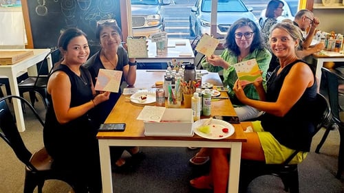Group of women sitting at a table showing finished painting projects