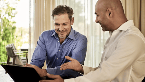 Two men in formal attire engage in a friendly discussion, smiling as they look at a laptop. Sunlit curtains provide a warm, inviting background.