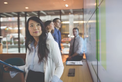A professional woman holding a tablet looks at a presentation screen, with colleagues blurred in the background, conveying a focused, collaborative atmosphere.