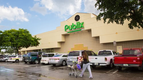 Woman pushing child in shopping cart past parked cars over wet pavement in front of Publix at Venetian Isle Shopping Center