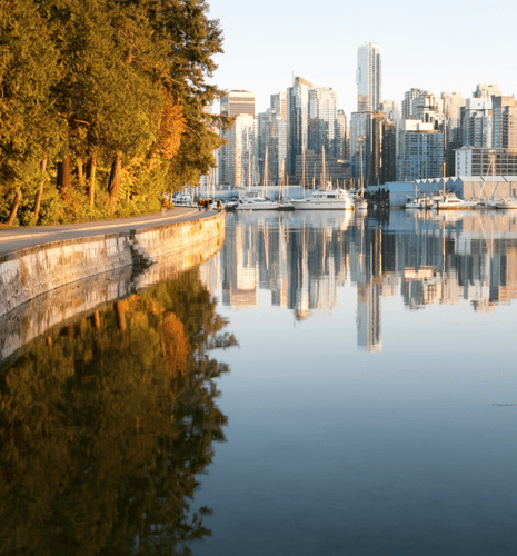 Calm water reflects a city skyline at sunset, bordered by a path lined with lush trees on the left, creating a serene and picturesque scene.