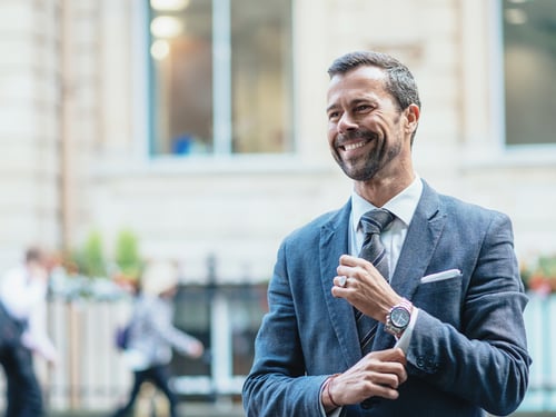 Smiling man in a suit adjusts his sleeve outdoors, exuding confidence. Blurred cityscape and pedestrians in the background convey busy urban life.