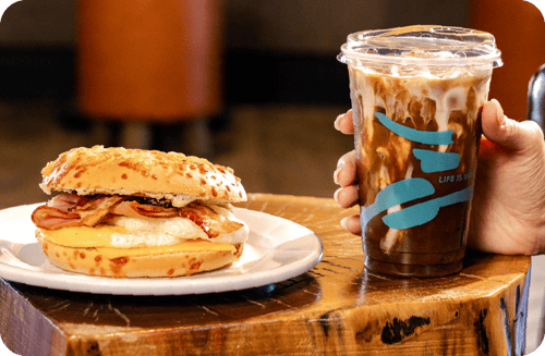 A hand holds a cold coffee drink in a plastic Caribou Coffee cup on a rustic table next to a breakfast sandwich.
