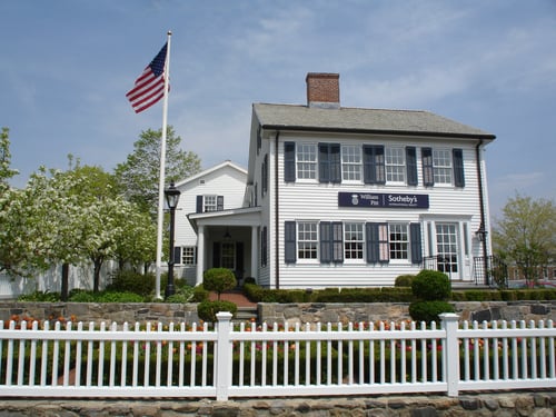White colonial-style building with black shutters housing William Pitt Sotheby's International Realty in New Canaan, Connecticut, featuring an American flag and a white picket fence.