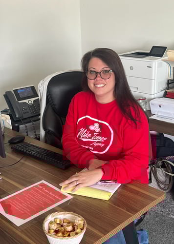 Smiling woman in red sweatshirt, seated at a wooden office desk with computer, phone, and documents. A bowl of candy adds a welcoming touch.