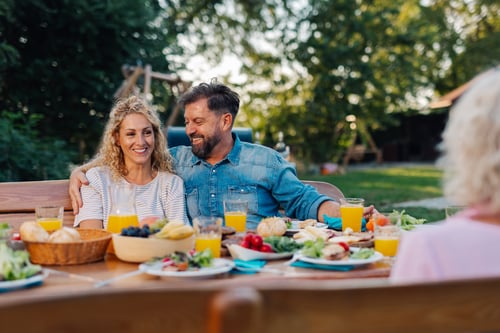 A couple smiles warmly at an outdoor brunch table filled with vibrant salads, fruits, and orange juice, set under lush trees. The scene exudes joy and relaxation.