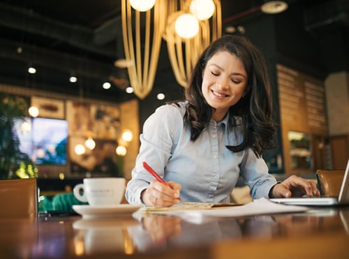 Woman at a coffee shop writing and on a laptop
