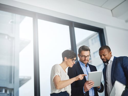 Three professionals collaborate, discussing something on a tablet in a bright, modern office. They appear focused and engaged in teamwork.