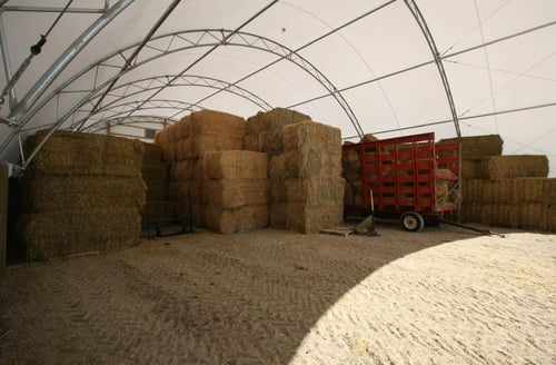 Stacks of hay bales and a red wagon stored inside a large Pilot Buildings fabric-covered agricultural structure.