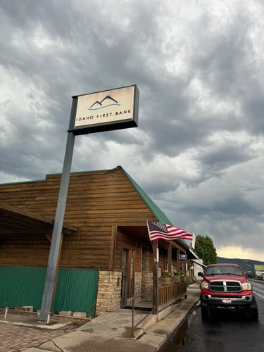 The exterior of an "Idaho First Bank" building with a rustic wooden facade and a green metal roof under a dramatic, cloudy sky. A "IDAHO FIRST BANK" sign is on a pole, and an American flag is displayed near the entrance. A red pickup truck is parked on the wet street in front of the bank.