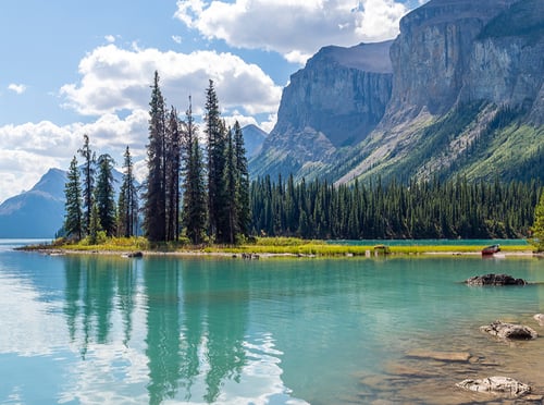 A serene lake scene with turquoise water reflecting tall evergreen trees and rocky mountains. Fluffy clouds scatter across the blue sky, evoking tranquility.