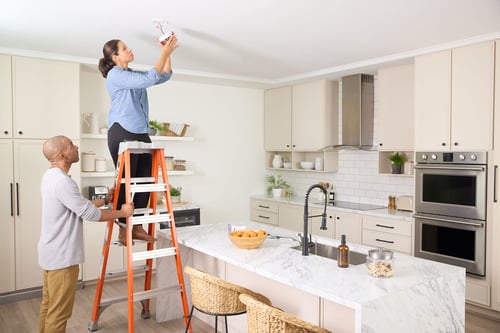 A woman installs a smoke alarm while her husband holds the ladder for safety.