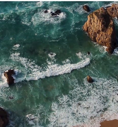 Overhead shot of waves crashing into rocks jutting out of the ocean.
