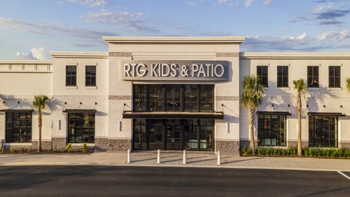 The main entrance of the RTG Kids & Patio store featuring bold signage, black-framed windows, and a palm-lined pathway on a sunny day.