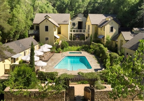 Bird's-eye view of yellow buildings around a pool area with rectangular pool, in-ground hot tub on the far end, and lounge chairs with umbrellas, all surrounded by trees.