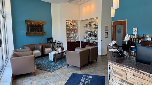 Clean, wide-angle interior view of the reception lobby at Hand & Stone Spa in South Loop, Chicago, showing plush leather guest armchairs, an immaculate check-in desk, a signature water wall, and fully stocked professional skincare retail displays.