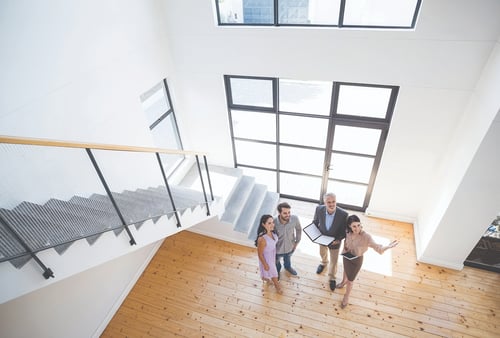 Aerial view of three people with a real estate agent inside a bright, spacious room with a wooden floor and large windows, suggesting a property tour.