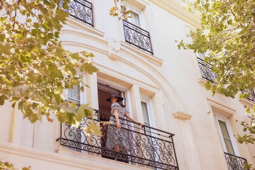 Woman with her back to the camera, holding on to a black steel railing outside an open balcony doorway on a beige building.