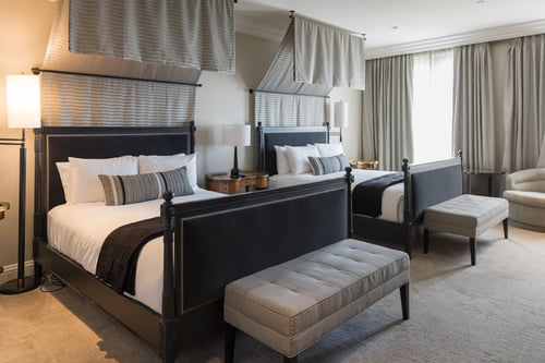 Two beds in a hotel room with a gray bench at each foot, the room adorned with various gray drapery and simple lamps.