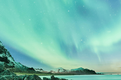 Aurora borealis streaking across starry sky above snow-capped mountains and turquoise water.