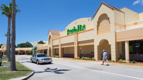Palm trees to the left of a silver car passing the front of Publix as a person walks by