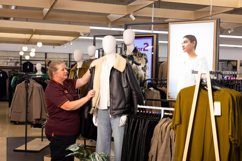 A Sainsbury's colleague adjusts a mannequin dressed in a black leather jacket, light shirt, and blue jeans inside a Sainsbury's store.