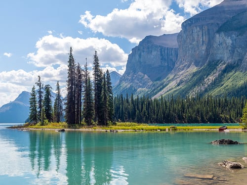 A serene lake scene with turquoise water reflecting tall evergreen trees and rocky mountains. Fluffy clouds scatter across the blue sky, evoking tranquility.