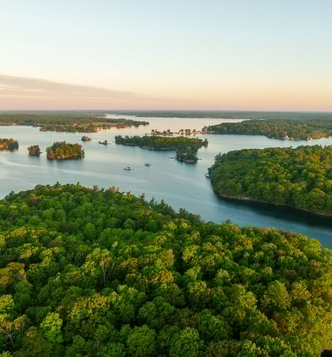 Aerial view of a serene lake with lush green forested islands under a clear sky. Boats dot the peaceful water, evoking tranquility and natural beauty.