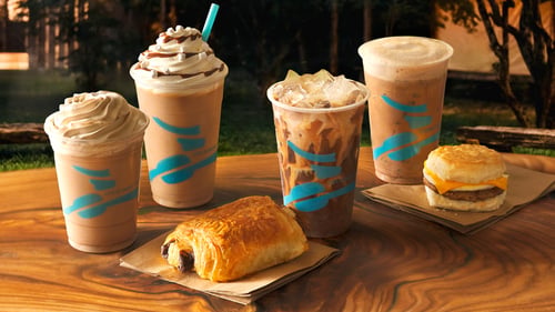 An assortment of iced and blended Caribou Coffee drinks with whipped cream on a wooden table, alongside a breakfast sandwich and croissant.