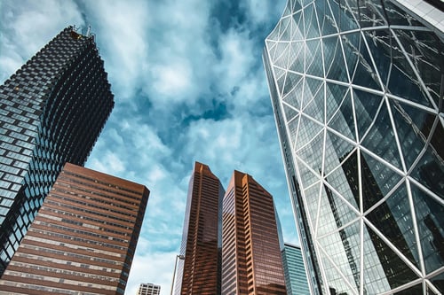 Calgary downtown skyline featuring modern skyscrapers including the distinctive Bow Tower with its diagonal lattice facade, viewed from street level on a winter day with dramatic cloudy skies.