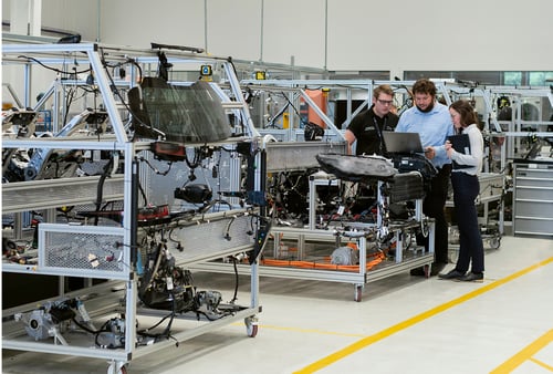 Three employees examine engine components and vehicle parts on workstations in an automotive manufacturing facility.